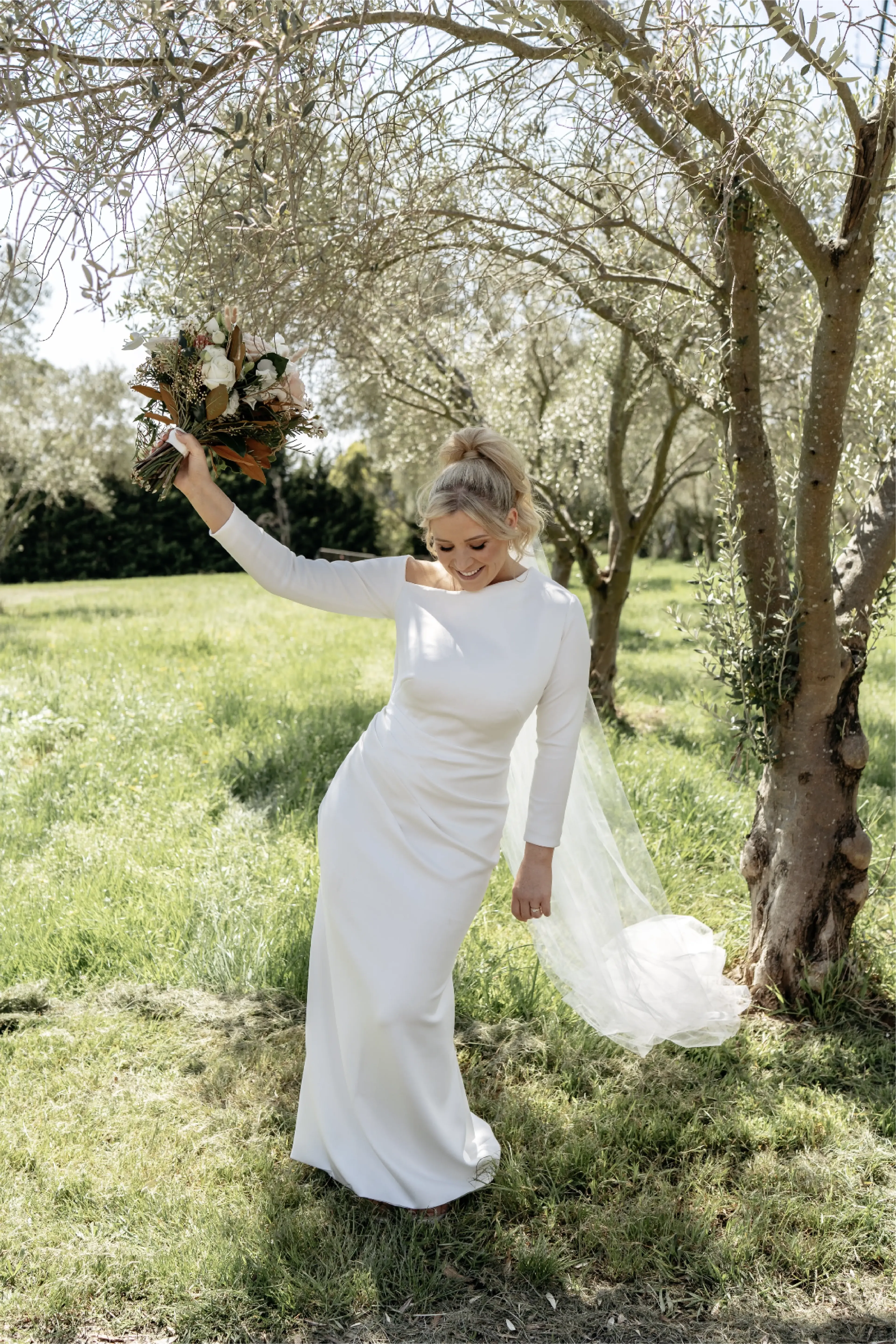 Bride standing under a tree wearing a high-neckline wedding dress with three-quarter sleeves. A delicate headpiece trails behind her as she holds a bouquet of flowers.